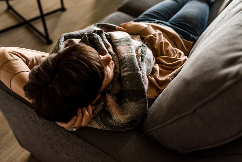 May include: A person is lying on a gray couch with a brown and gray blanket. They are wearing a brown shirt and blue jeans.
