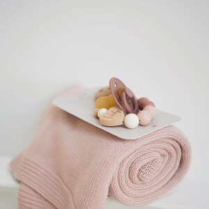Op de afbeelding: Een roze gebreide deken opgerold met een houten hartvormig bijtring met de naam "Vicky" erop gegraveerd. Het speelgoed is bevestigd aan een siliconen speen en een touwtje met houten kralen in verschillende roze tinten en beige.