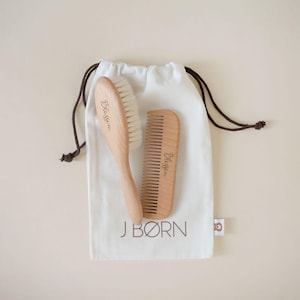 May include: A wooden baby brush and comb set with the word "Blissom" printed on each. The set is inside a white drawstring bag with the words "J BØRN" printed on it. The bag has brown drawstrings.