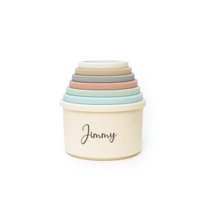 Peut inclure: Un jouet empilable avec une base crème et des anneaux aux couleurs pastel. Les anneaux sont dans les tons beige, gris, rose et bleu. Le nom "Jimmy" est écrit en cursive sur la base. Ce jouet est conçu pour les jeunes enfants.