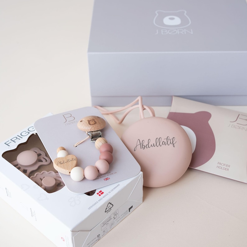 Op de afbeelding: Een cadeauset voor een baby met een roze en witte siliconen speen, een houten bijtring met een hartvormige hanger en een roze siliconen speenhouder met de naam "Abdullatif" erop gegraveerd.