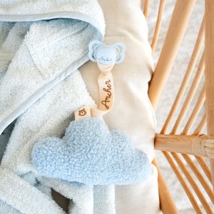 May include: A baby blue pacifier with the name "Archer" printed on it, attached to a beige ribbon with the name "Archer" printed on it. A fluffy blue cloud-shaped toy is attached to the ribbon. A light blue hooded towel is in the background.