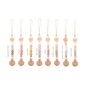 Op de afbeelding: Acht houten en siliconen baby-bijtjes met gepersonaliseerde hartcharms. Elke bijtje heeft een andere kleurencombinatie van siliconen kralen en een houten clip. De kleuren omvatten houtmarmot & blush, biscuit, kamille, lila & roze, pioenroos, vanille & room, poederblauw & room en salie & room.