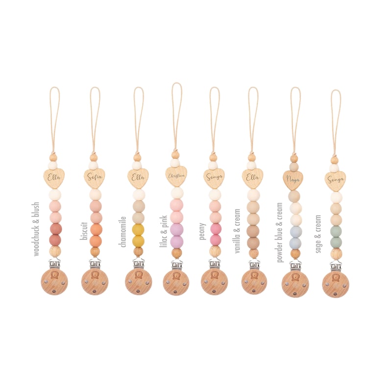 Op de afbeelding: Acht houten en siliconen baby-bijtjes met gepersonaliseerde hartcharms. Elke bijtje heeft een andere kleurencombinatie van siliconen kralen en een houten clip. De kleuren omvatten houtmarmot & blush, biscuit, kamille, lila & roze, pioenroos, vanille & room, poederblauw & room en salie & room.