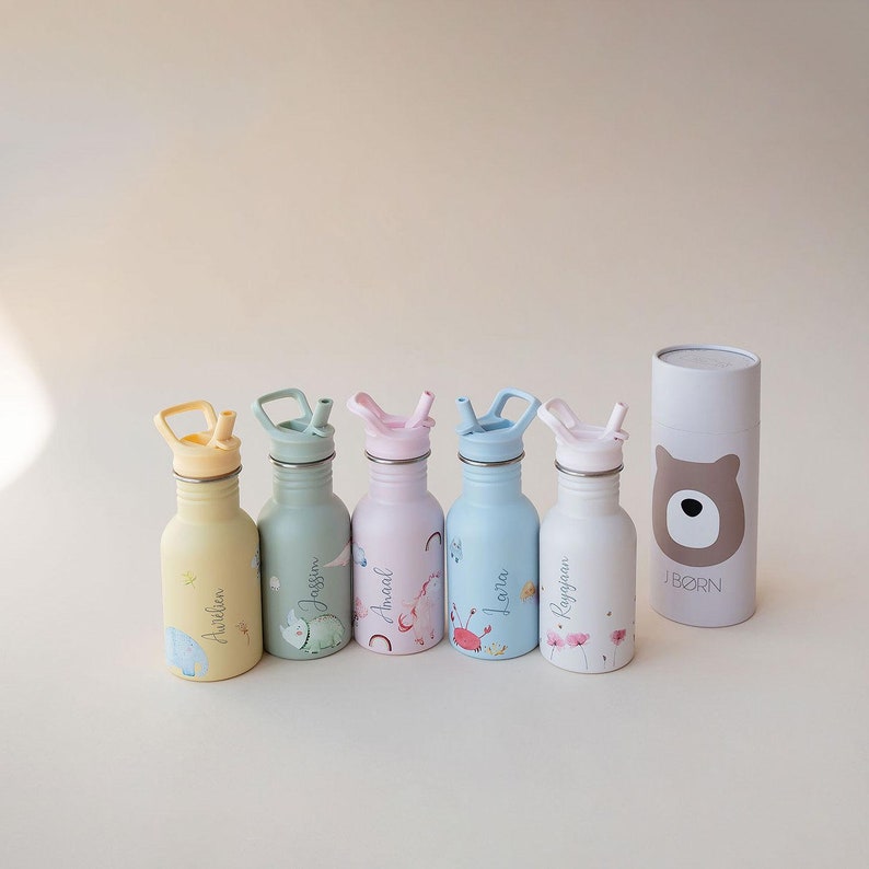 May include: Five colorful stainless steel water bottles with straw lids and personalized names. The bottles are decorated with floral and animal illustrations. The bottles are arranged in a row on a light beige surface. A white tube with a brown bear face and the word "J.BORN" is in the background.
