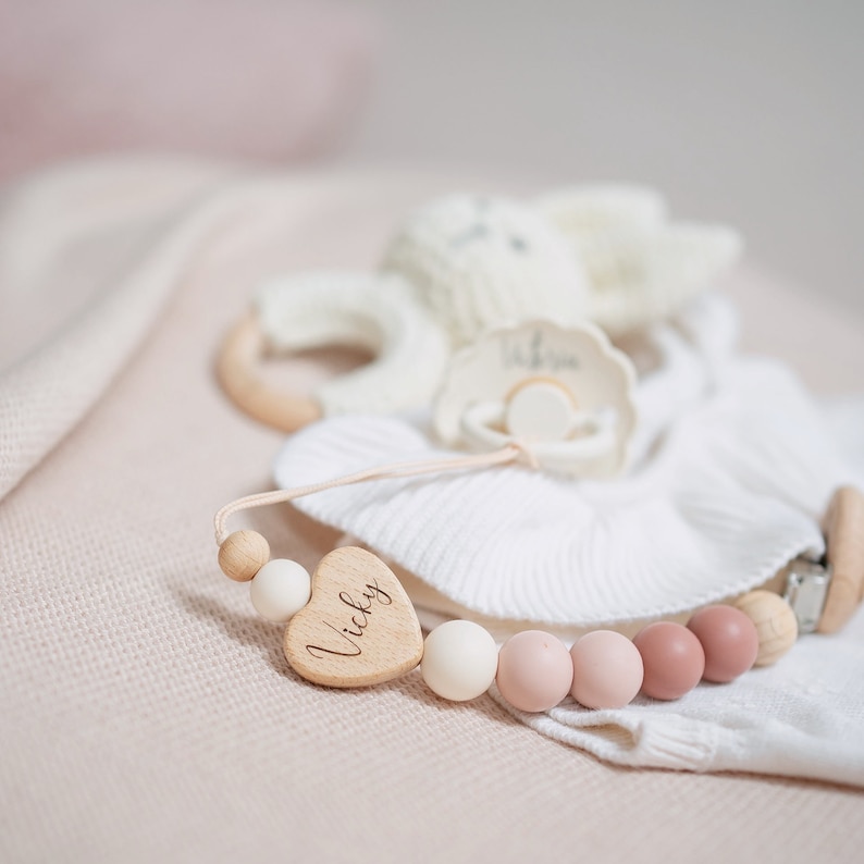 Op de afbeelding: Een houten hartvormige baby-bijtring met de naam "Vicky" erop gegraveerd. De bijtring is bevestigd aan een touwtje met houten en siliconen kralen in roze en witte tinten. De bijtring ligt op een wit gebreid dekentje.