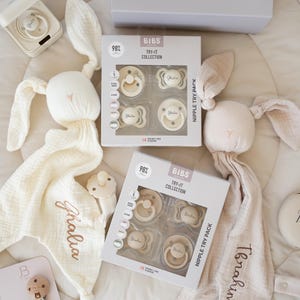 May include: An assortment of baby items in cream and beige tones. The image features two plush bunny security blankets with embroidered names, pacifiers, and boxed sets of pacifiers labeled "TRY-IT COLLECTION". The pacifiers are in various shapes and sizes.
