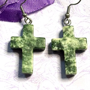May include: A pair of cross-shaped earrings. Each earring features a green stone cross with a speckled pattern, suspended from a silver-colored hook. The earrings are displayed against a purple fabric and a patterned background.