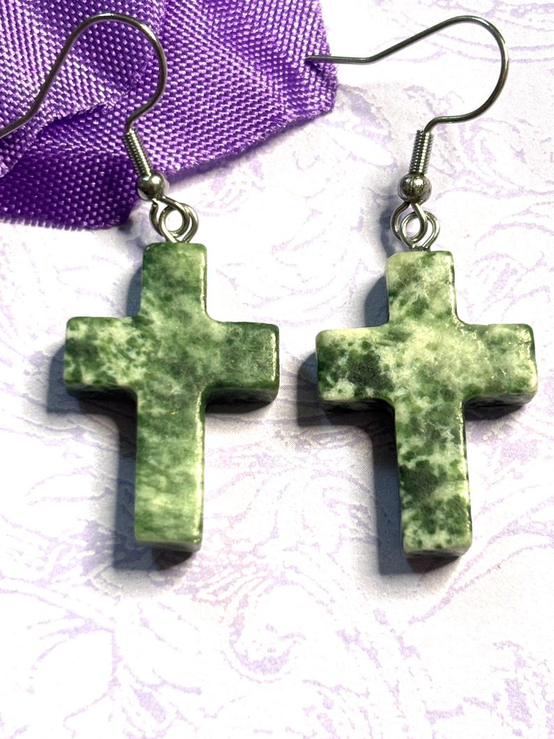 May include: A pair of cross-shaped earrings. Each earring features a green stone cross with a speckled pattern, suspended from a silver-colored hook. The earrings are displayed against a purple fabric and a patterned background.