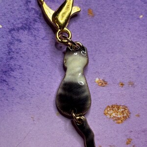 May include: A gold-colored lobster clasp with a charm of a cat silhouette. The cat charm is black and white with a gold-colored tail. The charm is attached to the clasp with a small gold-colored ring.
