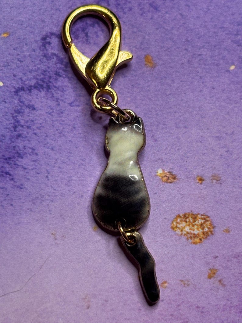May include: A gold-colored lobster clasp with a charm of a cat silhouette. The cat charm is black and white with a gold-colored tail. The charm is attached to the clasp with a small gold-colored ring.