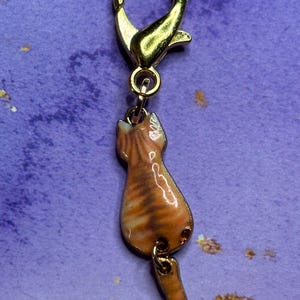 May include: A gold-colored charm featuring a tabby cat design. The charm is attached to a gold-colored clasp. The cat charm is orange and brown with a glossy finish. The cat is depicted from the back, with its tail hanging down.