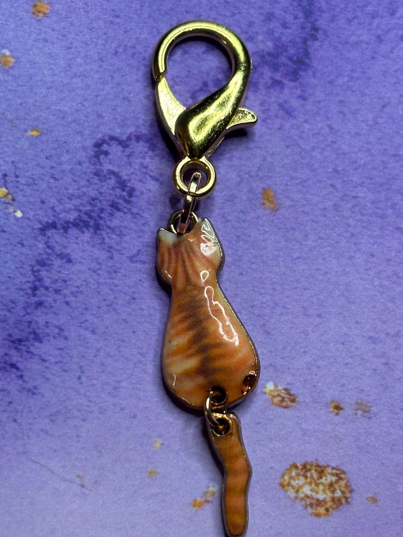 May include: A gold-colored charm featuring a tabby cat design. The charm is attached to a gold-colored clasp. The cat charm is orange and brown with a glossy finish. The cat is depicted from the back, with its tail hanging down.