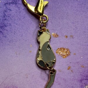 May include: A gold-colored metal cat charm with a lobster clasp. The cat charm is enamel-filled with a gray, white, and black design. The charm is attached to a gold-colored metal clasp, suitable for a keychain or zipper pull.