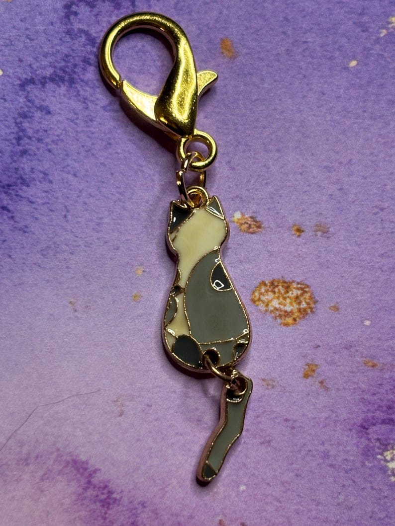 May include: A gold-colored metal cat charm with a lobster clasp. The cat charm is enamel-filled with a gray, white, and black design. The charm is attached to a gold-colored metal clasp, suitable for a keychain or zipper pull.
