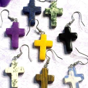 May include: A collection of cross-shaped earrings in various colors and materials. The earrings include purple, yellow, black, white, blue, and brown crosses, each with a unique texture and pattern. The earrings are suspended from silver-colored ear wires.