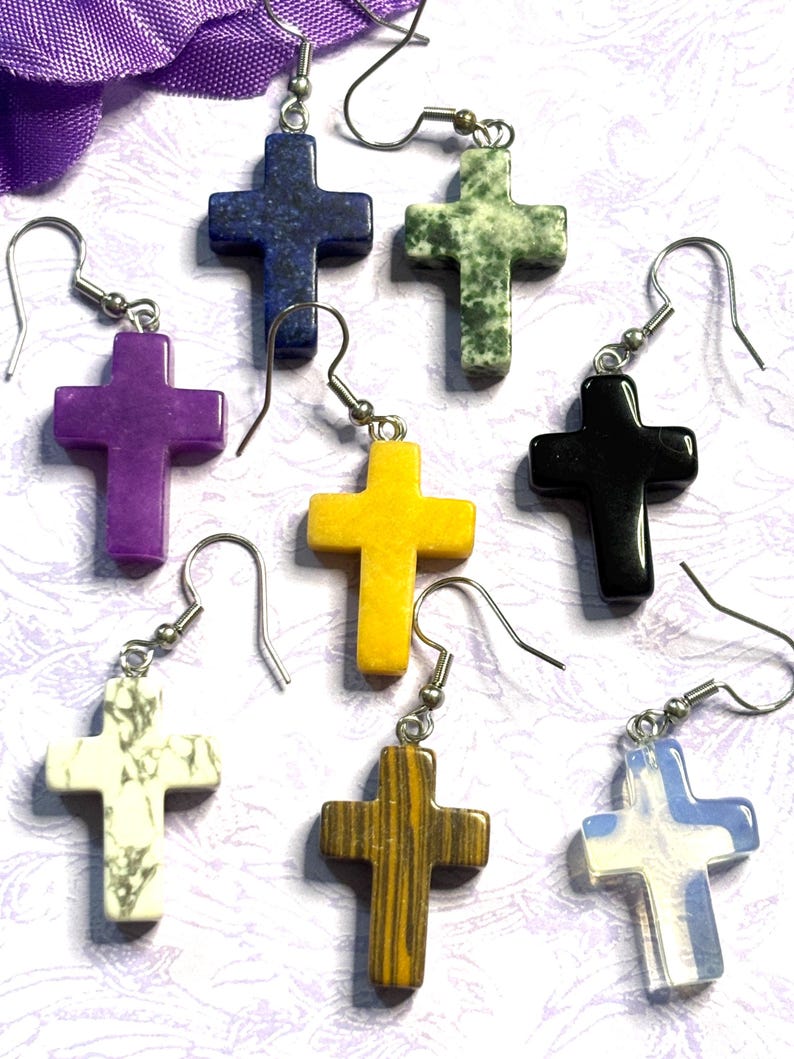 May include: A collection of cross-shaped earrings in various colors and materials. The earrings include purple, yellow, black, white, blue, and brown crosses, each with a unique texture and pattern. The earrings are suspended from silver-colored ear wires.