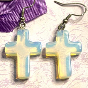 May include: A pair of cross-shaped earrings with a pearlescent, opalescent appearance. The earrings have silver-colored hooks and are displayed against a purple fabric and a patterned background. The crosses have a translucent quality.