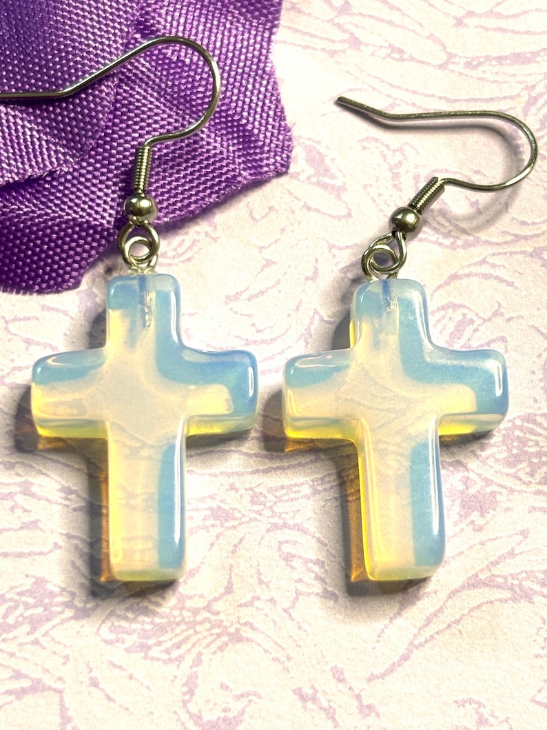 May include: A pair of cross-shaped earrings with a pearlescent, opalescent appearance. The earrings have silver-colored hooks and are displayed against a purple fabric and a patterned background. The crosses have a translucent quality.
