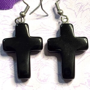 May include: A pair of black cross earrings with silver-colored hooks. The crosses are smooth and glossy, with a simple, classic design. The earrings are displayed against a light purple and white background.