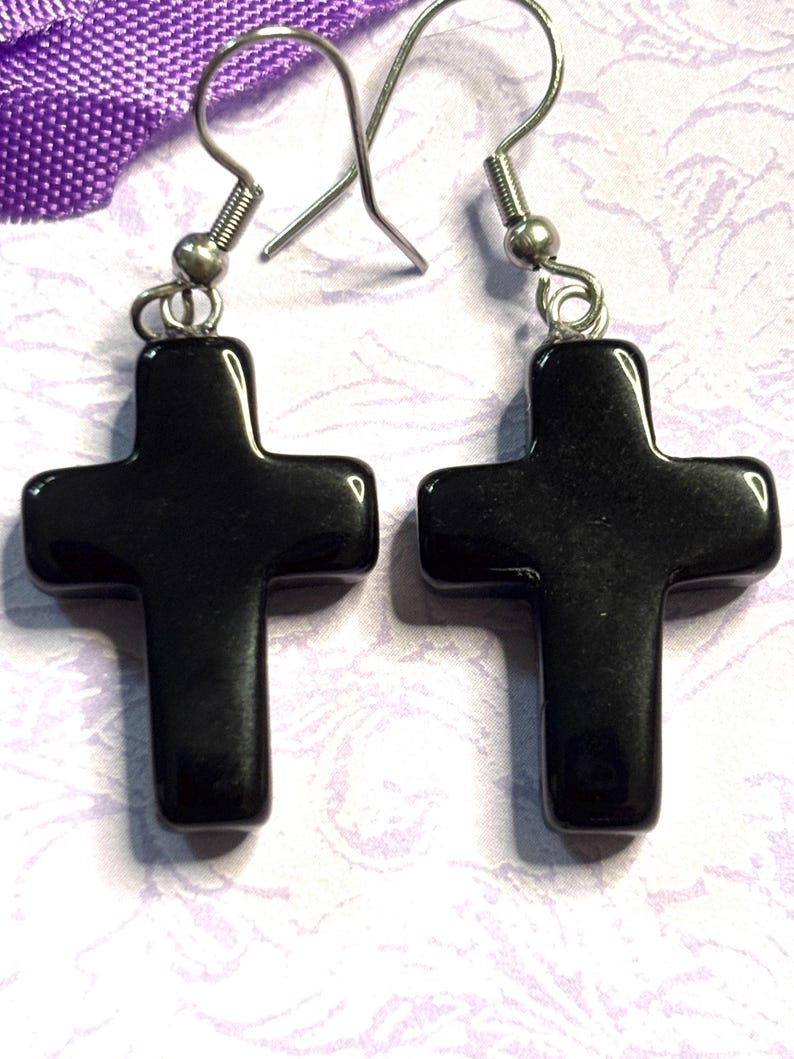 May include: A pair of black cross earrings with silver-colored hooks. The crosses are smooth and glossy, with a simple, classic design. The earrings are displayed against a light purple and white background.