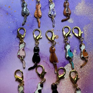 May include: A collection of cat-shaped charms with gold-colored clasps. The charms feature various cat designs, including tabby, calico, and solid colors, with detailed enamel work. The background is a purple and white marbled pattern.