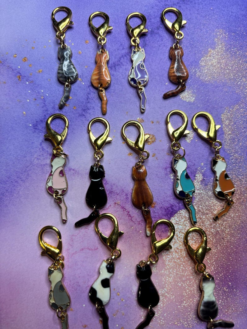 May include: A collection of cat-shaped charms with gold-colored clasps. The charms feature various cat designs, including tabby, calico, and solid colors, with detailed enamel work. The background is a purple and white marbled pattern.