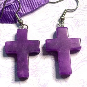 May include: A pair of purple cross earrings. Each earring features a polished, cross-shaped pendant suspended from a silver-colored fish hook. The earrings are displayed against a light background with a purple fabric.