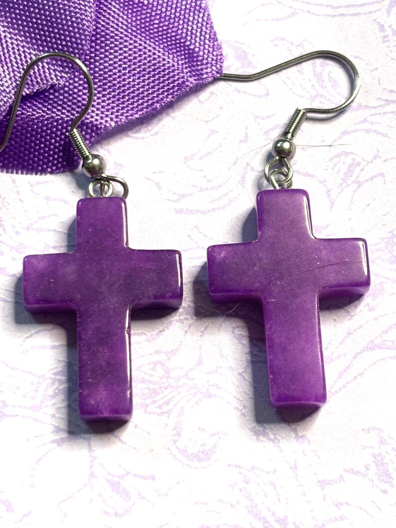 May include: A pair of purple cross earrings. Each earring features a polished, cross-shaped pendant suspended from a silver-colored fish hook. The earrings are displayed against a light background with a purple fabric.