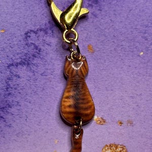 May include: A gold-colored lobster clasp with a small, brown tabby cat charm. The cat charm is detailed with stripes and a curled tail, hanging from a small chain. The clasp is attached to a purple background.
