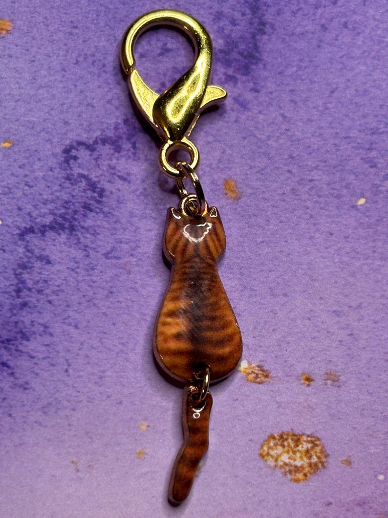 May include: A gold-colored lobster clasp with a small, brown tabby cat charm. The cat charm is detailed with stripes and a curled tail, hanging from a small chain. The clasp is attached to a purple background.