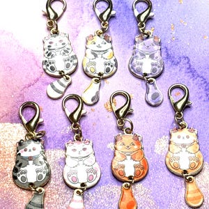May include: A collection of seven cat-shaped charms with gold-colored clasps. Each charm features a different color scheme, including black and white, orange, and purple. Each cat charm has a dangling teardrop-shaped charm attached.