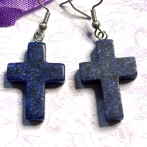 May include: A pair of earrings featuring dark blue cross pendants with silver-colored hooks. The crosses have a speckled pattern and are displayed against a soft, patterned background. The earrings are a religious accessory.