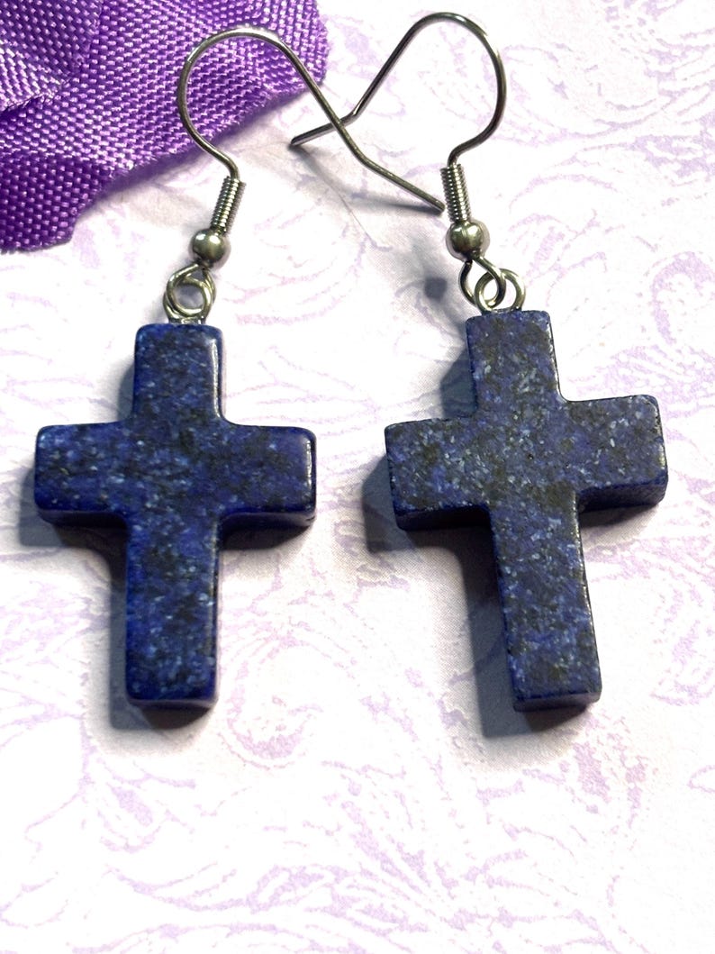 May include: A pair of earrings featuring dark blue cross pendants with silver-colored hooks. The crosses have a speckled pattern and are displayed against a soft, patterned background. The earrings are a religious accessory.