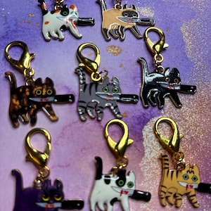 May include: A collection of cat-shaped charms with gold-colored clasps. Each charm features a different colored cat holding a black knife in its mouth. The cats are in various colors, including white, black, orange, and grey.
