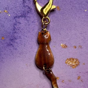 May include: A gold-colored metal clasp with a brown cat charm. The cat charm is brown with a white-tipped tail. The charm is attached to the clasp with a small gold ring. The background is a purple color.
