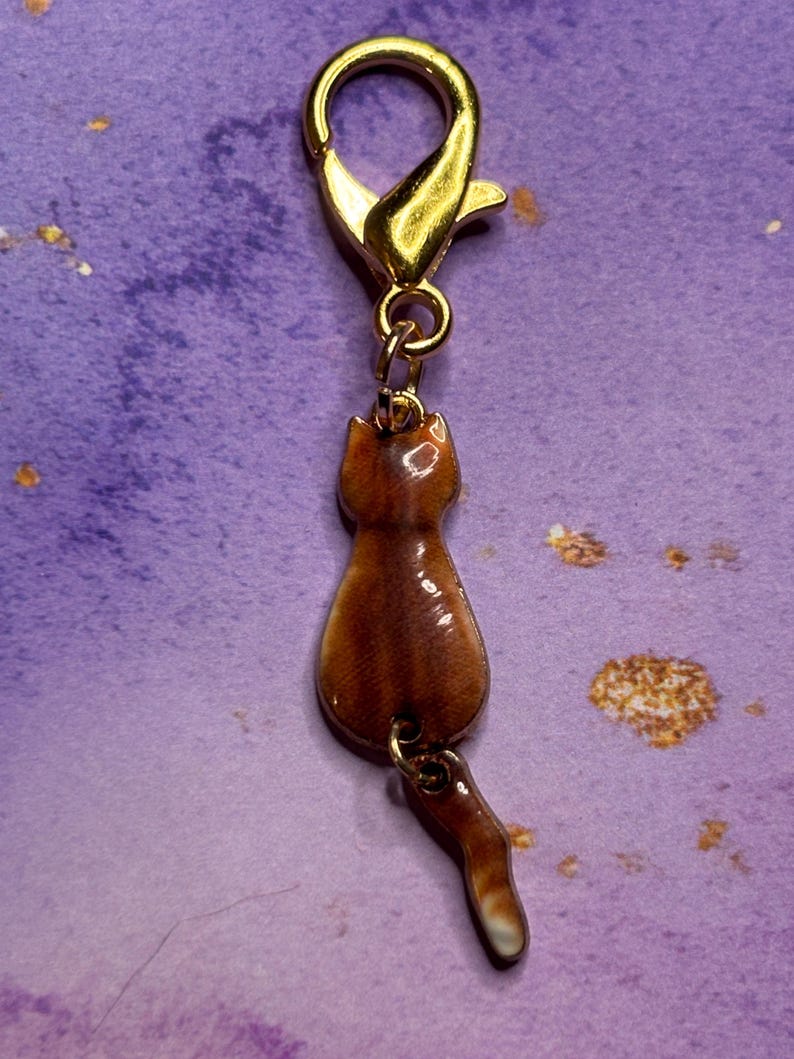 May include: A gold-colored metal clasp with a brown cat charm. The cat charm is brown with a white-tipped tail. The charm is attached to the clasp with a small gold ring. The background is a purple color.