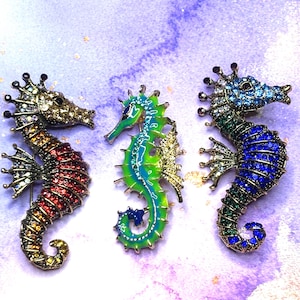 May include: Three seahorse brooches in red, green, and blue. Each brooch is embellished with rhinestones and has a detailed design. The seahorses have a gold-tone metal base and are set against a purple and white background.
