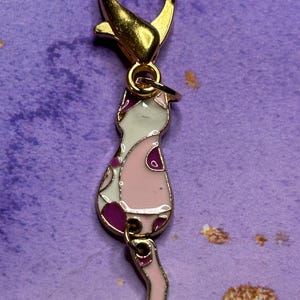 May include: A gold-colored metal cat charm with a lobster clasp. The cat charm is pink, white, and purple with a patterned design. The charm is designed to be attached to a keychain or necklace.