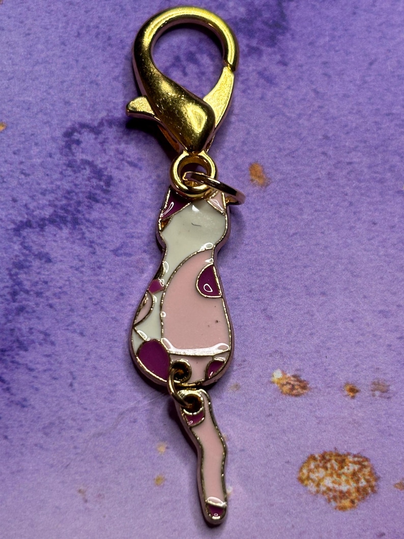 May include: A gold-colored metal cat charm with a lobster clasp. The cat charm is pink, white, and purple with a patterned design. The charm is designed to be attached to a keychain or necklace.