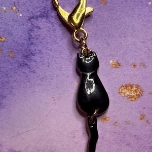 May include: A gold-colored lobster clasp with a small, black cat charm. The cat charm is glossy and has a detailed silhouette, including a tail. The background is a textured purple with gold flecks.