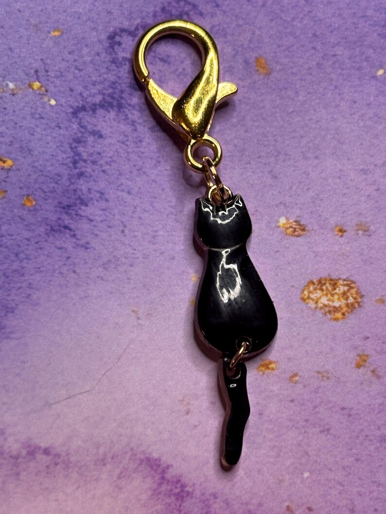 May include: A gold-colored lobster clasp with a small, black cat charm. The cat charm is glossy and has a detailed silhouette, including a tail. The background is a textured purple with gold flecks.