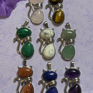 May include: A collection of cat-shaped pendants with various gemstones. The pendants feature a silver-colored metal frame and include stones like rose quartz, tiger's eye, malachite, and amethyst. The cat design has a head and body, with a tail.