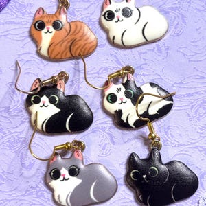 May include: Six pairs of cat-shaped earrings with gold-colored hooks. The cats are in various colors, including orange tabby, white, black, gray, and black and white. Each cat has a cute, cartoonish design with large eyes and a smiling face.