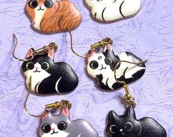May include: Six pairs of cat-shaped earrings with gold-colored hooks. The cats are in various colors, including orange tabby, white, black, gray, and black and white. Each cat has a cute, cartoonish design with large eyes and a smiling face.