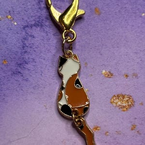 May include: A gold-colored lobster clasp with a charm of a cat. The cat charm is white, brown, and black, with a tail hanging down. The charm is attached to the clasp with a small gold ring.