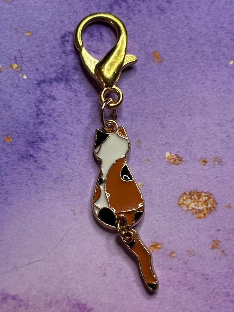 May include: A gold-colored lobster clasp with a charm of a cat. The cat charm is white, brown, and black, with a tail hanging down. The charm is attached to the clasp with a small gold ring.