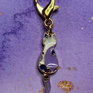 May include: A gold-colored metal cat charm with a lobster clasp. The cat charm is purple, white, and navy blue with gold-colored trim. The charm is a decorative accessory.