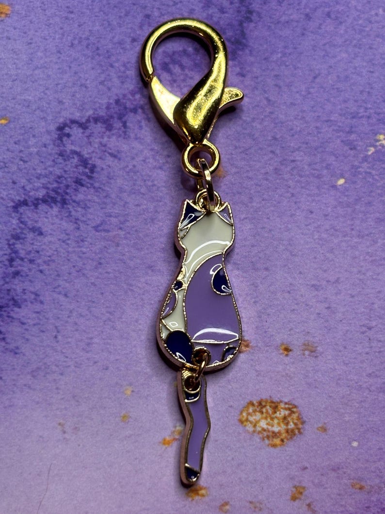 May include: A gold-colored metal cat charm with a lobster clasp. The cat charm is purple, white, and navy blue with gold-colored trim. The charm is a decorative accessory.