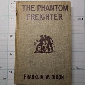 Może przedstawiać: Antyczna książka w miękkiej oprawie zatytułowana "The Phantom Freighter" autorstwa Franklina W. Dixona. Okładka przedstawia brązowe tło z sylwetką dwóch mężczyzn biegnących przez pole wysokiej trawy.